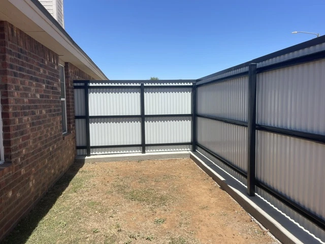 Modern welded steel privacy fence showing back of ribbed panels and black frame, built on a continuous concrete footing to enclose a backyard beside a brick house.