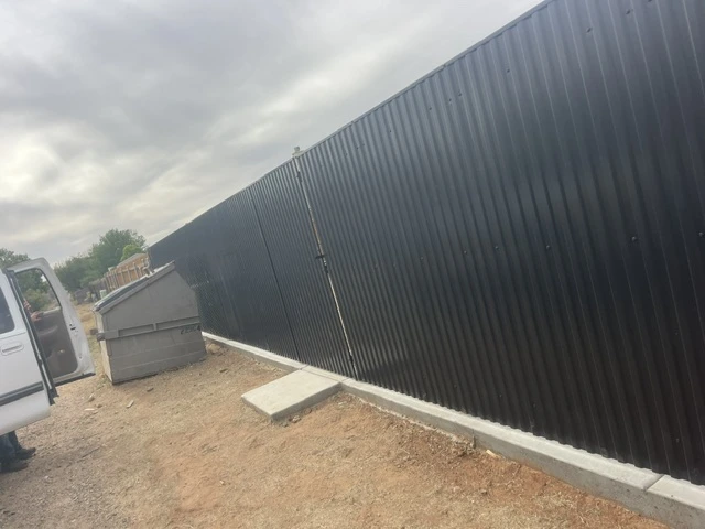 Custom black metal privacy fence with vertical ribbed panels and steel posts installed along a concrete footing at a site in Texas.